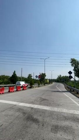 The video shows a clear sky with power lines stretching across it. In the foreground, a grey asphalt road is visible, divided by white lines. Red barriers are placed along the left side of the road. Beyond the barriers, a white van and some trees can be seen. A road sign with circular symbols is also present. The road curves to the right in the distance, with streetlights lining its edge. On the right side of the road, a metal guardrail is installed, and trees and foliage are visible. A diamond-shaped black and yellow sign is placed on a pole on the right side of the road. The scene appears to be a highway or a major road on a sunny day.