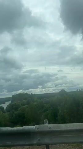 The camera view is from a moving vehicle, looking over a metal safety railing towards a wide river and a distant landscape of trees under an overcast sky. The vehicle is on a bridge, and the river flows horizontally across the frame. The riverbanks are lined with dense green foliage. The sky is filled with grey clouds, creating a subdued light.
