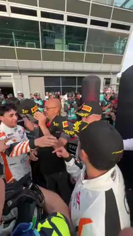 The video captures a lively celebratory moment at a racetrack, likely after a race, followed by a brief view of the track.

**Subjects:** A group of approximately 15-20 people are gathered tightly. Among them are several race car drivers, dressed in white racing suits with black and orange details. One suit prominently displays an "Audi" logo and "SANLORENZO" text. The drivers are wearing black baseball caps adorned with a yellow laurel wreath design and a "Pirelli" logo, indicating a victory or podium finish. Other individuals, possibly team members or officials, are present. One man in a black polo shirt has "1ST" printed on his sleeve, and another wears a similar shirt with a laurel wreath design. A man in a white collared shirt is also actively participating in the interaction.

**Activity:** The people are engaged in a celebratory exchange, with smiles and gestures. The men in casual attire are seen placing the "Pirelli" caps onto the heads of the race car drivers, signifying congratulations. The atmosphere is joyous and exciting.

**Notable details:** The initial scene is set in what appears to be a pit lane or garage area, with a modern building featuring large windows in the background. The lighting suggests it is daytime. The second part of the video briefly pans to show a green and white race car, bearing the text "BIOCYCLE," on the track. In the distance, a large yellow archway with the "PIRELLI" brand name is visible over the track.

**Visible text:** "1ST" is visible on a black polo shirt. "AUDI" and "SANLORENZO" are on a white racing suit. "Pirelli" logos are on the caps and the distant archway. "BIOCYCLE" is on the green and white race car.

**Location:** Monza, Italy.