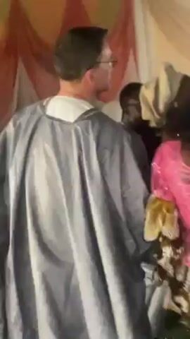 Here is a concise description of the video:

The video shows a man in a gray, loose-fitting robe surrounded by several women in colorful African dress. The event appears to be a celebration, with dancing and music playing in the background. The man and women are dancing together.