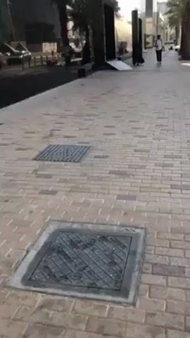 The video shows a pedestrian view of a paved sidewalk. The sidewalk is made of grey bricks. There are two metal drain covers in the sidewalk. The sidewalk is next to a street. A building with mirrored glass windows is seen on the left. The windows reflect the buildings across the street. People are walking on the sidewalk. There are trees and benches on the sidewalk. The sky is overcast.