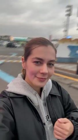 A woman is taking a selfie video outside on an overcast day, possibly in a parking lot. She wears a black leather jacket over a grey hoodie. The background features a large, lit-up Christmas tree, a carousel adorned with flowers, and a building in the distance. She turns the camera to showcase the Christmas decorations and smiles at the camera.
