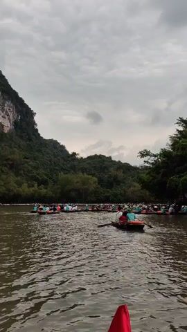 The video shows a river filled with boats, many of which are packed closely together with passengers wearing blue shirts or hats. The camera appears to be positioned low, possibly from another boat, with a red object visible in the foreground. On either side of the river are lush green landscapes, including a forested hill on the left and trees lining the right bank. The sky is overcast, suggesting a cloudy day. The water is calm with gentle ripples, reflecting the overcast sky.
