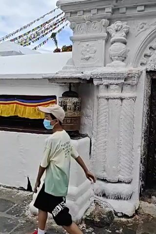 A man wearing a white cap and a green and white t-shirt is seen from the back, walking towards a white stupa. He is wearing black shorts and white socks with red trim. Beside him, attached to a white pedestal, is a prayer wheel. The prayer wheel is cylindrical and appears to be made of metal with intricate carvings. In the background, prayer flags are strung across the top of a white structure, and beyond that, the golden dome of a large stupa is visible against a cloudy sky. The stupa itself is white with decorative carvings and features an ornate entrance. There are also orange garlands draped over the white structure. Another person, wearing a white headscarf and a blue jacket, is partially visible in front of the ornate entrance. The overall scene suggests a religious or spiritual site, likely in a mountainous region given the prayer flags and the architectural style.