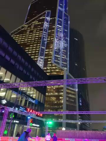 The video shows an ice skating rink under the lights in front of tall buildings at night. Several people are skating. Overhead lighting rigs provide illumination. The rink is surrounded by short walls bearing promotional graphics. The buildings in the background are modern skyscrapers with illuminated windows. The sky is overcast.
