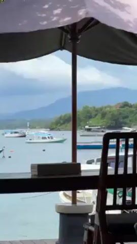The video showcases an outdoor restaurant or cafe with a view of a harbor or bay. The scene is shot from within the partially enclosed restaurant space, looking out towards the water. Several boats are visible in the water, with land and trees in the background. The foreground shows tables and chairs under umbrellas, typical of an outdoor dining setting. A potted plant is on one of the tables. There's a sense of a relaxed, possibly tourist-oriented atmosphere. The camera pans slightly, showing more tables and chairs in the restaurant.