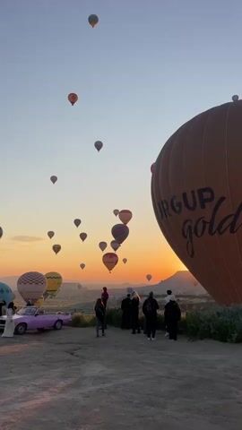 The video captures a picturesque scene of hot air balloons ascending into a clear sky during either sunrise or sunset in Göreme, Turkey. A large, orange hot air balloon, prominently displaying the text "ÜRGÜP gold," dominates the right foreground. A group of people are gathered on a dusty, rocky ground, observing the spectacle. Some are looking up at the sky, while others are engaging with their surroundings.

To the left, a distinct vintage-style pink convertible car is parked, adding a unique aesthetic to the scene. Beyond the immediate foreground, numerous other hot air balloons of varying sizes and colors dot the vast sky, some already high up, others closer to the horizon where the sun casts a warm, golden glow across the landscape. The background features the characteristic hilly terrain and rock formations of Cappadocia. The overall atmosphere appears calm and appreciative as people witness the iconic hot air balloon event.