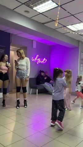The camera captures a scene within what appears to be a dance studio or event space with a vibrant purple and white color scheme. In the background, a glowing neon sign reads "why not?" illuminating a group of people casually seated on a gray couch. The room is decorated with string lights hanging from the ceiling.

In the foreground, several individuals are standing and conversing. Three adult women are wearing dance outfits, including high-heeled boots, short bottoms, and knee pads. They seem to be engaged in conversation, with one holding a beverage cup and another looking at her phone.

Interspersed among the adults are several young children, both boys and girls, who are moving and dancing, suggesting an informal or practice setting. One of the boys is wearing a green shirt and gray shorts. The girls are dressed in casual attire.

The studio has a clean tiled floor and a large poster visible on the wall near the entrance. Overall, the atmosphere seems relaxed and lively, blending a mix of adults and children in a dance-related environment.