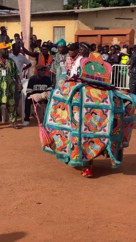 The video captures a lively outdoor event. The focal point is a person in an elaborate, colorful costume, adorned with beads and intricate patterns. The costume is wide and layered, with a distinctive hat featuring a green dragon-like figure and the word "OGIRI" prominently displayed. The wearer's feet, clad in red and yellow shoes, are visible as they move.

Surrounding the costumed individual are numerous onlookers, some seated on white chairs under a white tent, others standing. The environment appears to be an open, dusty area, possibly a village square or a community gathering space, with buildings visible in the background. Many people are dressed in traditional African attire, adding to the festive atmosphere. Some individuals in the crowd are holding cameras and phones, recording the spectacle. The overall mood seems to be celebratory and engaging.