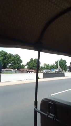 The video is taken from inside a vehicle, likely a tuk-tuk, looking outwards. The interior of the vehicle's roof is visible in the foreground, with a patterned fabric. A metal pole runs vertically down the center of the frame. Outside, there is a road with two lanes, separated by a concrete barrier with black and white markings. On the other side of the barrier, there is a side of the road with some buildings, trees, and a few people. Traffic is visible on the road, including a motorcycle, a car, and another tuk-tuk. The weather appears to be overcast. The perspective suggests the vehicle is in motion.