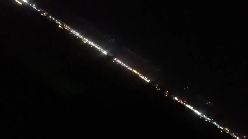From a high angle, the video depicts a line of lights that may be vehicles or city lights, illuminated at night. The perspective is diagonal, with the lights stretching across the frame. Some yellow runway lights are seen in the foreground. The surrounding environment appears dark and undefined.