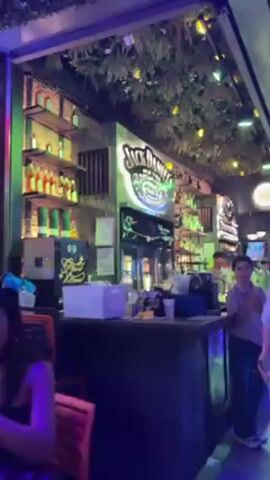 The video shows the interior of a crowded bar at night. Patrons sit at tables, and a screen shows a soccer match. The bar has neon signs that say "Koko Hostel". The lighting is colorful. People are wearing casual clothing.