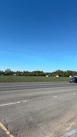 The video depicts a grey car traveling on a road on a sunny day. The sky is clear and blue, and a field with scattered buildings and trees is visible on the side of the road. The car is moving from right to left in the frame. Power lines can be seen overhead.