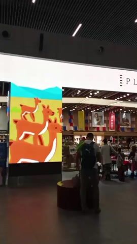 The video shows an indoor scene, possibly a shopping mall or airport terminal. A large LED screen displays an animated image of four deer stacked on top of each other against a yellow and blue background. In front of the screen, a man wearing a green backpack and gray pants stands with his back to the camera, looking towards a store. Behind him, other people are visible, some browsing merchandise within the store. The store's signage appears to say "PL..." and displays various banners with portraits. The overall environment is well-lit with a modern ceiling structure.