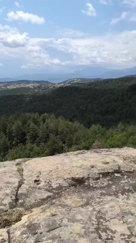 A video shows a panoramic view of a mountainous landscape with lush green forests. The camera pans across the scenery from a rocky overlook. The sky is partly cloudy with white and gray clouds. The foreground features large rocks and vegetation. A wooden fence is visible at some points, suggesting a hiking trail or viewpoint. The video captures the natural beauty of the mountainous terrain and the dense forest cover.