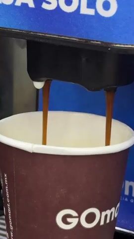 A close-up shot shows a brown paper cup with a white rim, branded with "GOmart" and "gomart.com.mx", receiving two steady streams of dark liquid from a black coffee machine dispenser. One of the dispenser spouts has a small white drip on it. The cup is being filled, with the liquid already covering the bottom. In the background, a blue surface with partial white text, "CA SOLO", is visible, suggesting a coffee machine or display for black coffee. The scene takes place at a GOmart location, likely a convenience store or gas station in San Buenaventura, Mexico.