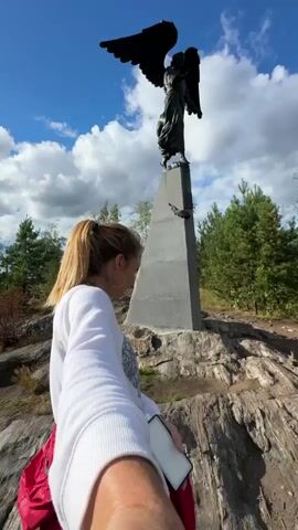 The video begins with a low-angle shot looking up at a dark angel statue on a tall, grey, pyramid-shaped monument against a blue sky with white clouds. In the foreground, a woman with blonde hair pulled back in a ponytail, wearing a white sweatshirt and a red bag on her left shoulder, is holding her right arm out as if taking a selfie. She is looking towards the monument. The camera then pans down to show the rocky terrain and moves to the right, revealing a man in a camouflage shirt and grey pants standing on the rocky ground. He is holding a phone in his right hand and looking down at it. The background shows green trees, a body of water, and a cloudy sky.