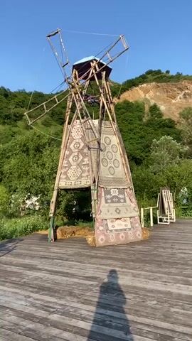 A windmill-like structure, adorned with rugs, stands on a wooden platform. The structure is made of wood and metal and has rotating blades at the top. The rugs, which have intricate patterns, are draped down the sides of the structure. The windmill is located in an outdoor setting with trees and a hill in the background. The sky is clear and blue. The sun is casting shadows on the wooden platform. There is no visible human activity.