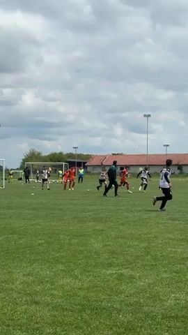 A soccer game is in progress on a green field. Young players in orange and black and white uniforms are running and kicking. A coach in dark clothing walks along the sideline. In the background are buildings, trees, light poles, and a cloudy sky.