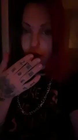 The video shows a woman with red hair, wearing a black shirt with a silver chain necklace, smoking a cigarette and blowing out smoke. She has tattoos on her hand. The background is dark and unclear. There is music playing.