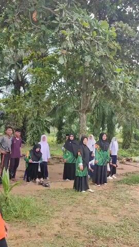 A group of young women, most wearing hijabs and green and black uniforms, are gathered outdoors in a park or field. One young woman in the foreground, wearing a black hijab and an orange shirt, appears to be in motion, possibly throwing something. Another young woman in the background, wearing a red shirt, stands near a target. The environment appears to be somewhat rural, with trees and vegetation visible. The scene suggests a sporting event or activity, possibly archery or a similar throwing game.