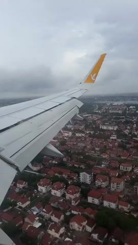 The video shows an aerial view from an airplane window looking out over a city. The wing of the airplane, with the Pegasus Airlines logo visible on the winglet, dominates the left side of the frame. Below, a dense urban landscape unfolds, characterized by numerous houses with red tile roofs. Trees are interspersed throughout the residential areas. In the distance, the city stretches out towards a hazy horizon, under a cloudy sky. The perspective suggests the aircraft is in the process of descending or ascending, as the city appears relatively close. No people are visible within the frame.