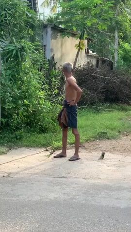 The video captures a street scene, likely in a village or rural area. In the foreground, a lean, shirtless young man stands with his hands on his hips, wearing a sarong tied around his waist and flip-flops. He is facing to his left, looking away from the camera. Behind him, lush green foliage and a simple building with a beige wall and a blue roof are visible. Further down the street, a small shop or stall is set up, displaying various products on shelves. A woman in red and white patterned pants and a gray t-shirt walks past the shop. To the right of the shop, a brown dog sits on the side of the road. In the immediate foreground, the hindquarters of a cow are visible, suggesting it is either walking or tethered nearby. The ground is a mixture of dirt and a paved road. The overall lighting suggests it is daytime, possibly late afternoon due to the soft light.