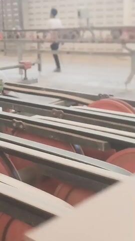 The video shows a manufacturing or processing facility. In the foreground, a conveyor belt system with red rollers and black belts is visible. The belts are in motion, suggesting an active process. In the background, two individuals are present, appearing to be workers. One person is wearing a light-colored shirt and dark pants, and the other is wearing a white shirt and dark pants. They are positioned near machinery and appear to be overseeing or participating in the production line. The environment is dusty, indicating a process that generates particulate matter, such as woodworking or material processing. The overall impression is of an industrial setting where materials are being moved and processed.