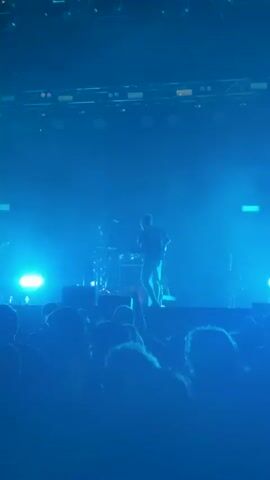 A concert crowd faces a dimly lit stage where a musician, dressed in a dark shirt and shorts, performs under bright blue and yellow lights. The stage features musical instruments, including drums and guitars, and a fog machine creates a hazy atmosphere. The audience, mostly out of focus, is visible from behind, with some individuals holding up their phones. The video pans across the scene, capturing the dynamic lighting and the energy of the performance.