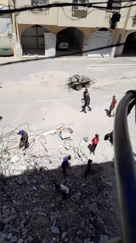 The video shows a high-angle view of a street covered with debris. Several people are walking on the street, carrying what appear to be water jugs. One person stumbles and drops their jug, spilling its contents on the ground. In the background, a small wagon is laden with twisted metal. There is a pile of rubble in the foreground. Buildings are visible in the background.