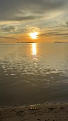 A vertical, medium shot captures a serene beach sunset. The sun, a brilliant orb, hangs low on the horizon, casting a golden reflection across the calm sea. Wispy clouds stretch across the sky, tinged with hues of orange and grey, suggesting the transition from day to night. In the distance, the silhouettes of islands emerge from the sea. The foreground features a sandy beach with gentle waves lapping at the shore. No people or specific actions are visible. The overall atmosphere is one of peaceful tranquility.