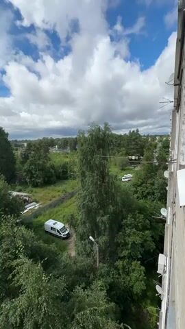 The video opens with a wide shot of a cloudy sky with patches of blue visible. A tall tree dominates the center of the frame, with a white van parked on a dirt path to the left of it. In the distance, there are more trees and some buildings. The camera then pans to the right, revealing more trees and the side of a building. The video then shifts to an interior shot of a kitchen. A stove with pots and pans on it is visible on the left. A toaster and other kitchen items are on the counter to the right.