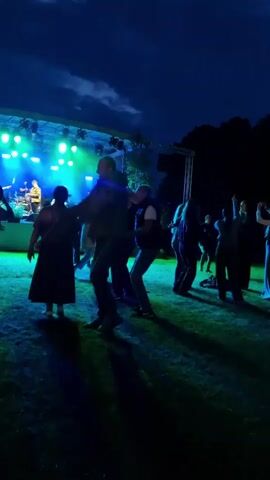 The video shows a group of people dancing on a grassy area in front of a stage at night. A band is performing on the stage, illuminated by blue and green lights. The sky is dark with some clouds. One person is being lifted in the air by another person in the foreground. Other people are dancing and interacting with each other. The overall atmosphere appears to be festive and energetic.