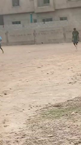 The video depicts a sandy, open area in what appears to be a residential neighborhood.  There are several people, mostly young men, present in the video.  Some are standing, some are sitting on a low brick wall, and others are walking.  The people are wearing casual clothing.  The environment consists of low-rise buildings, some of which have satellite dishes on them.  A car passes by on what seems to be a dirt road.  The ground is uneven, covered with sand and some sparse vegetation. The overall impression is of a casual, unpaved area where people are gathering or playing.