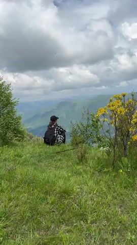 A lone individual wearing a plaid shirt and a black baseball cap is sitting on a grassy hilltop, facing away from the camera. They have a black backpack on their back. The person is looking out over a vast valley with rolling green hills and a river running through it. The sky is cloudy with some patches of blue. There are trees and yellow wildflowers in the foreground. The video is filmed from a medium distance, capturing the person and the expansive landscape.