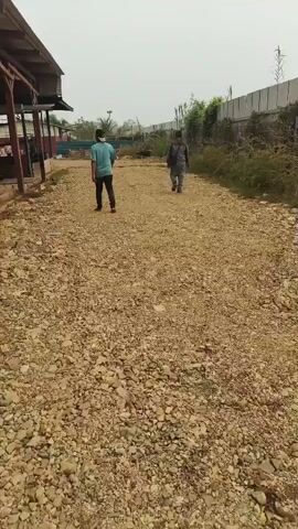The video shows two individuals walking away from the camera on a gravel path. The person in the foreground on the left wears a light blue t-shirt and dark pants, and appears to be wearing a face mask. The person in the background on the right wears a dark shirt and pants, and carries a backpack. To the left of the path is a structure with a brown roof and supporting columns, appearing to be an open-sided building or shelter. The ground is covered in loose gravel and some larger stones. To the right of the path is dense vegetation and a fence. The sky is overcast. The scene suggests an outdoor, possibly industrial or construction site, environment.