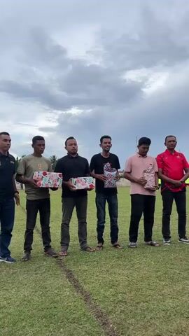 Six men stand in a line on a grassy field under a cloudy sky. They are all facing forward, towards the camera, and appear to be receiving or holding gifts. From left to right, the first man is wearing a black polo shirt with a logo on the left chest and blue jeans. The second man is wearing a green t-shirt and dark pants, holding a box wrapped in colorful patterned paper. The third man is wearing a black t-shirt and dark pants, also holding a similarly wrapped box. The fourth man is wearing a black t-shirt and blue jeans, holding a wrapped box. The fifth man is wearing a pink t-shirt and dark pants, holding a wrapped box. The sixth man is wearing a red polo shirt and dark pants. Another person, mostly out of frame, is standing to the right of the sixth man, appearing to be taking a photo. There are white lines on the grass in front of the men, suggesting it is a sports field. In the background, there are some trees and structures.