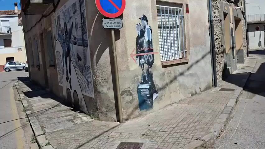 The video captures a street scene in Salt, Spain, likely during the day. The camera pans across a weathered building corner adorned with street art. On the left, a large mural depicts two figures in a stylized, black-and-white sketch, seemingly walking or dancing, with elements of a cityscape and a bench in the background. To the right of the mural, a different piece of graffiti shows a silhouette of a figure in black and blue, holding what appears to be a bottle, with red lettering and an arrow pointing left. Above the graffiti, a street sign is visible. The sign has a blue circle with a red diagonal line through it, indicating a prohibition, likely related to parking or stopping, and below it, a rectangular sign with text in what appears to be Catalan. The text on the rectangular sign reads "Dia 13" and then "de 10:00 a 13:00" followed by "Excepte festius," suggesting a time restriction on the 13th of the month, excluding holidays.

The street itself is paved with square tiles and asphalt, with a curb and a gutter. A silver car is parked on the left side of the street. In the background, residential buildings with balconies and windows are visible, suggesting an urban or semi-urban environment. The lighting indicates it's a sunny day. The overall impression is a snapshot of a street with artistic interventions, offering a glimpse into the local culture and environment of Salt. The emotions conveyed are neutral, focusing on observation of the scene.