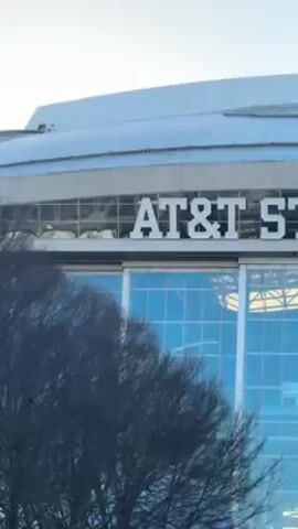 The video shows the exterior of AT&T Stadium, home of the Dallas Cowboys. The stadium has a large sign with its name, "AT&T STADIUM," displayed prominently. The building has a glass facade and a rounded roof. A man walks in front of the stadium. There are some trees and metal barricades in the foreground. The sky is clear and blue.