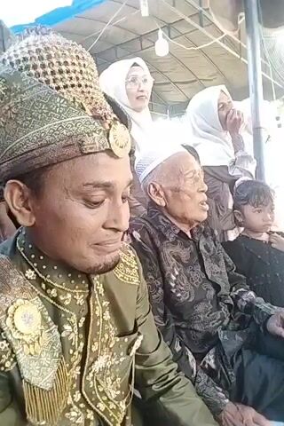 The video shows a close-up of a man in traditional attire, possibly a groom, wearing an ornate, green and gold headdress and a matching elaborately decorated jacket. His face is slightly turned to the side, and he appears to be dabbing his eye with a white tissue. Behind him, an elderly man in a dark patterned shirt and a white cap is visible, looking forward and speaking. To the right, a young girl in dark clothing is also present, looking off-camera. In the background, two women wearing hijabs are visible, one of them looking towards the front with an expression of concern. The setting appears to be an outdoor event, possibly a wedding or a traditional ceremony, with a tent-like structure overhead and natural light. The overall mood seems to be emotional, given the man's action of dabbing his eye.