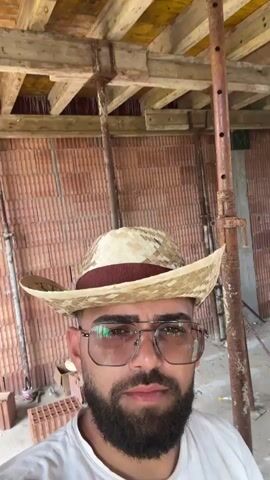 A selfie video shows a bearded man wearing a straw hat and glasses. He is standing inside a building under construction, with brick walls, wooden beams, and scaffolding visible in the background. The lighting is natural, suggesting it is daytime. The man is looking directly at the camera, and his expression is neutral. There are no other people or significant actions taking place in the video.