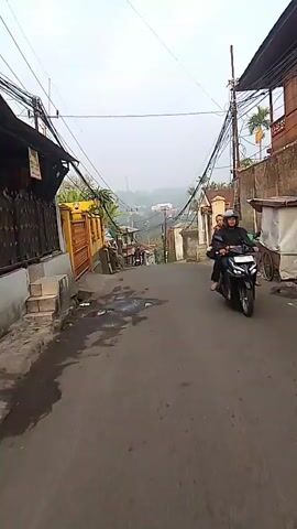 The video shows a downward sloping street in a densely populated area. The street is paved and wet in places, suggesting recent rain. Numerous overhead wires crisscross the street, supported by poles on either side. Buildings line both sides of the street, with some appearing to be homes and others small businesses. There are several parked motorcycles and a few people visible on foot and riding motorcycles. The overall impression is of a well-trafficked, somewhat cluttered, and potentially hilly urban environment. The weather appears overcast.