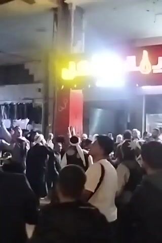 A crowd of people gathered in an urban setting, likely a street or plaza, are engaged in what appears to be a celebration or performance. The majority of individuals are men, and several are wearing traditional headdresses. One man is prominent, dressed in a vest and sporting a white headdress, gesturing animatedly with his hands, seemingly leading the group. The surrounding area is illuminated by bright, diffused artificial light, as well as neon signs and storefronts. The overall atmosphere suggests a lively, festive event.