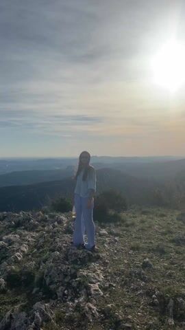 A young woman stands on a rocky hilltop, bathed in bright sunlight. She is wearing light blue, loose-fitting clothing and sunglasses. In her hand, she holds a bottle. Behind her, a mountainous landscape stretches into the distance under a partly cloudy sky. The sun is high and prominent, creating a strong backlight.