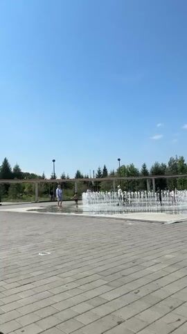 The video shows a sunny day in a park. In the foreground, a paved area with large square tiles. In the midground, a large, shallow water feature with multiple jets of water spraying upwards, creating a mist. Several people, including children and adults, are interacting with the water feature, some walking through it and others standing nearby. In the background, there is a treeline and a clear blue sky with a few wispy clouds. A covered walkway with support columns runs along the edge of the paved area and the treeline.