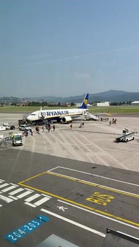A Ryanair airplane is parked on the tarmac at an airport, with passengers boarding via a mobile staircase. Several vehicles, including a baggage tug and what appears to be a small bus or van, are positioned around the aircraft. People are visible on the tarmac, some walking towards the plane and others near the boarding stairs. The scene is set against a backdrop of distant hills and a clear sky. The ground is marked with yellow and white lines, indicating a runway or taxiway.