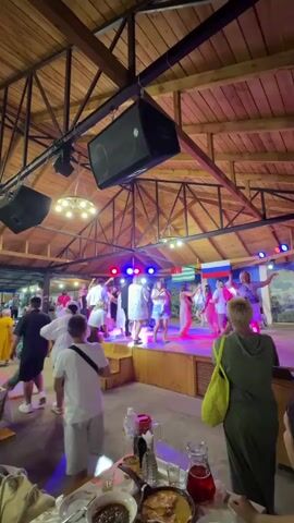 The video shows an indoor venue with a wooden roof structure. A raised stage is set up at one end where a group of people in white clothing are dancing, illuminated by colored stage lights. Russian flags are visible behind the performers. The venue appears to be a restaurant or banquet hall, with tables and chairs set up in the foreground and mid-ground. Several people are seated at the tables, while others are standing and watching the performance. A young boy in a white t-shirt and grey shorts is in the foreground, facing the stage. A woman in a green dress with a yellow bag is also in the foreground, facing away from the camera. Speakers are mounted on the ceiling structure above the stage. The overall atmosphere appears to be festive.