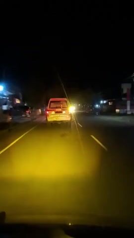 The video displays a first-person perspective from inside a vehicle driving at night. The road ahead is illuminated by strong yellow headlights from the filming vehicle. A white van with red markings on its rear is visible directly in front, driving in the same direction. Several oncoming vehicles with bright white headlights are seen approaching on the opposite side of the road. The environment is dark with some distant lights barely visible on the sides of the road.