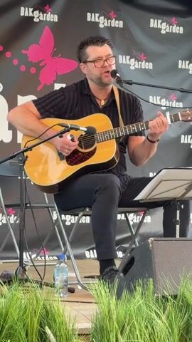 A man with glasses and a black collared shirt sits on a chair, playing an acoustic guitar and singing into a microphone. He is on a stage, with a banner behind him that says "BAKGARDEN" with a pink butterfly graphic. There are microphones, a music stand with sheet music, a water bottle, and speakers on the stage. In the foreground, there is green grass.