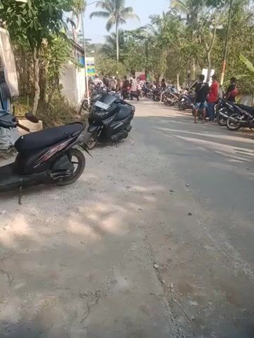 The video displays a street scene with several motorcycles parked along the side. The road appears to be made of asphalt or concrete and is cracked in places. Lush green trees and foliage line the left side of the road. A group of people are gathered on the left side, some standing and others appear to be sitting on or near the motorcycles. The sun is shining, casting shadows on the road, suggesting it is daytime. The overall impression is that of a busy area with parked vehicles and people.